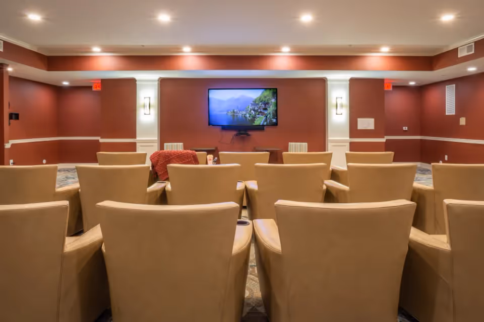 Rows of tan armchairs arranged theater-style facing a wall-mounted TV in a cozy screening room with red walls and recessed lighting.