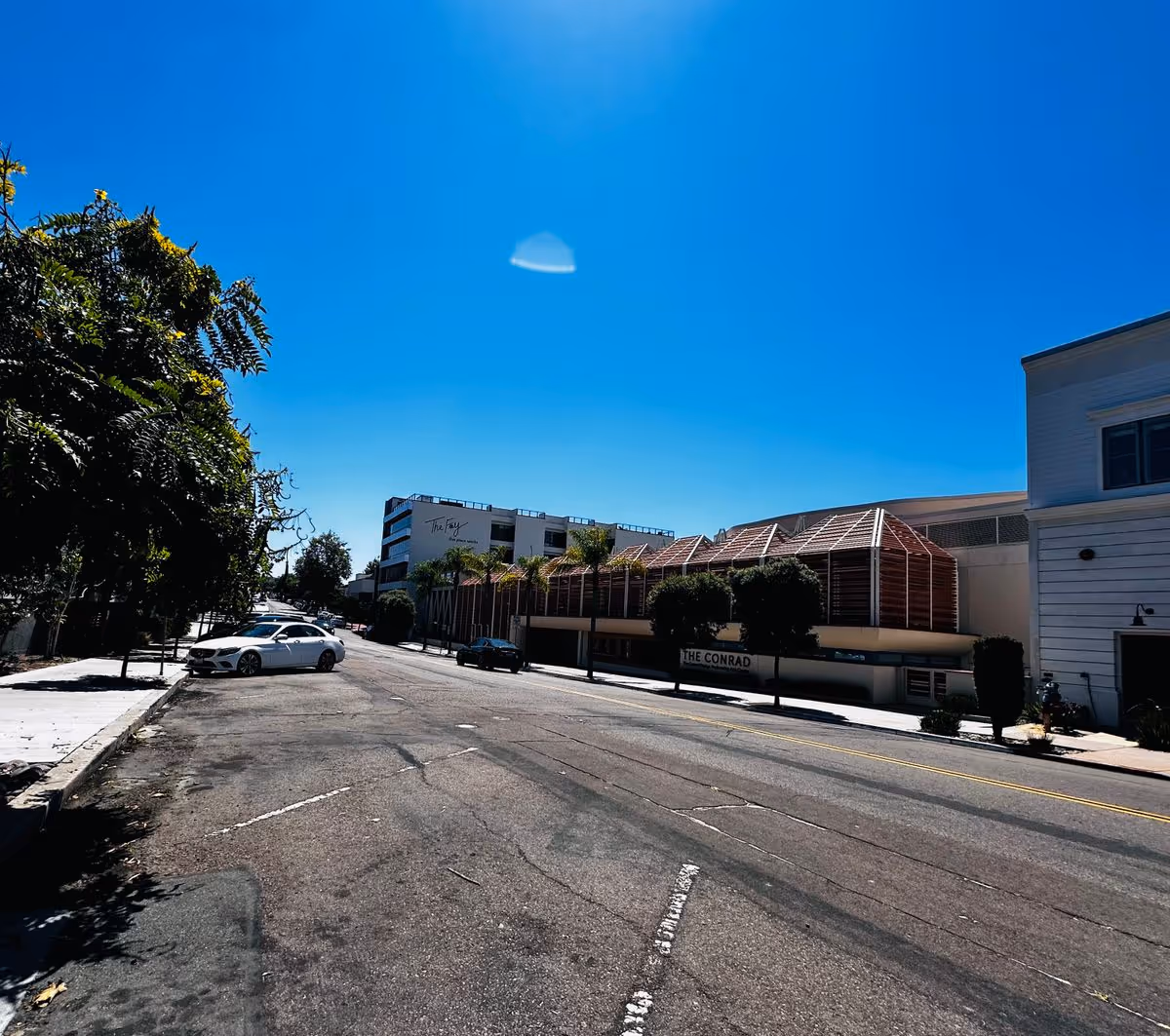 Sunlit empty street with parked cars, trees, and low-rise buildings under a clear blue sky.