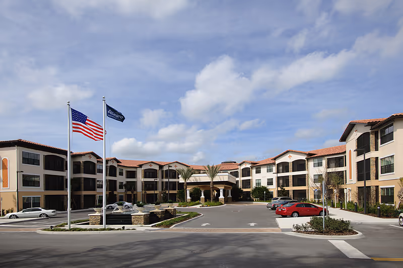 Front exterior of a Mediterranean-style senior living building with flags, a circular driveway, and parked cars under a blue sky.