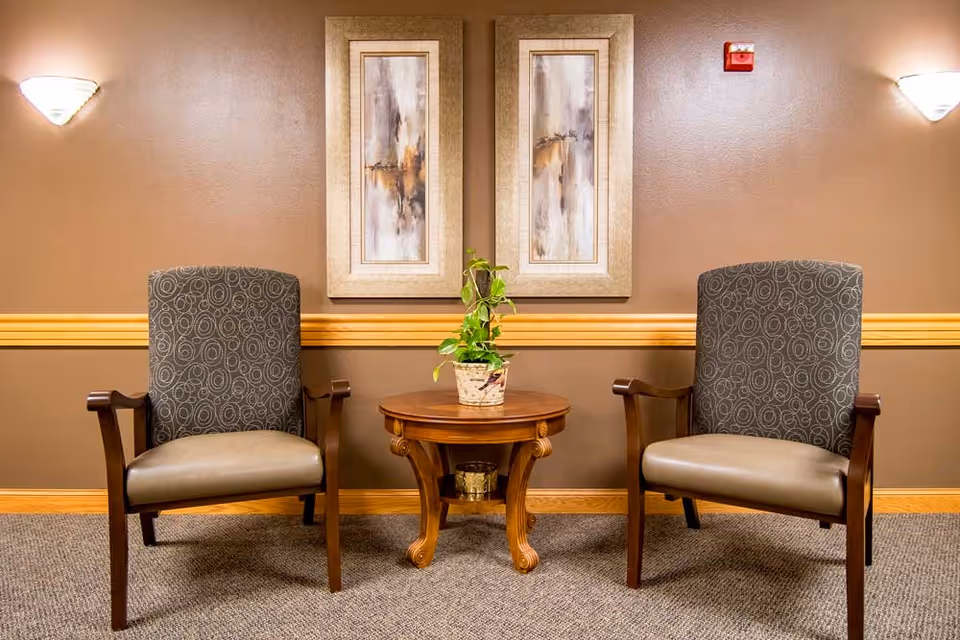 Two patterned armchairs with wooden arms are placed on either side of a small round wooden table with a potted plant on top, set against a brown wall with a wooden chair rail and two framed abstract paintings above the table. Wall sconces provide soft lighting on both sides.