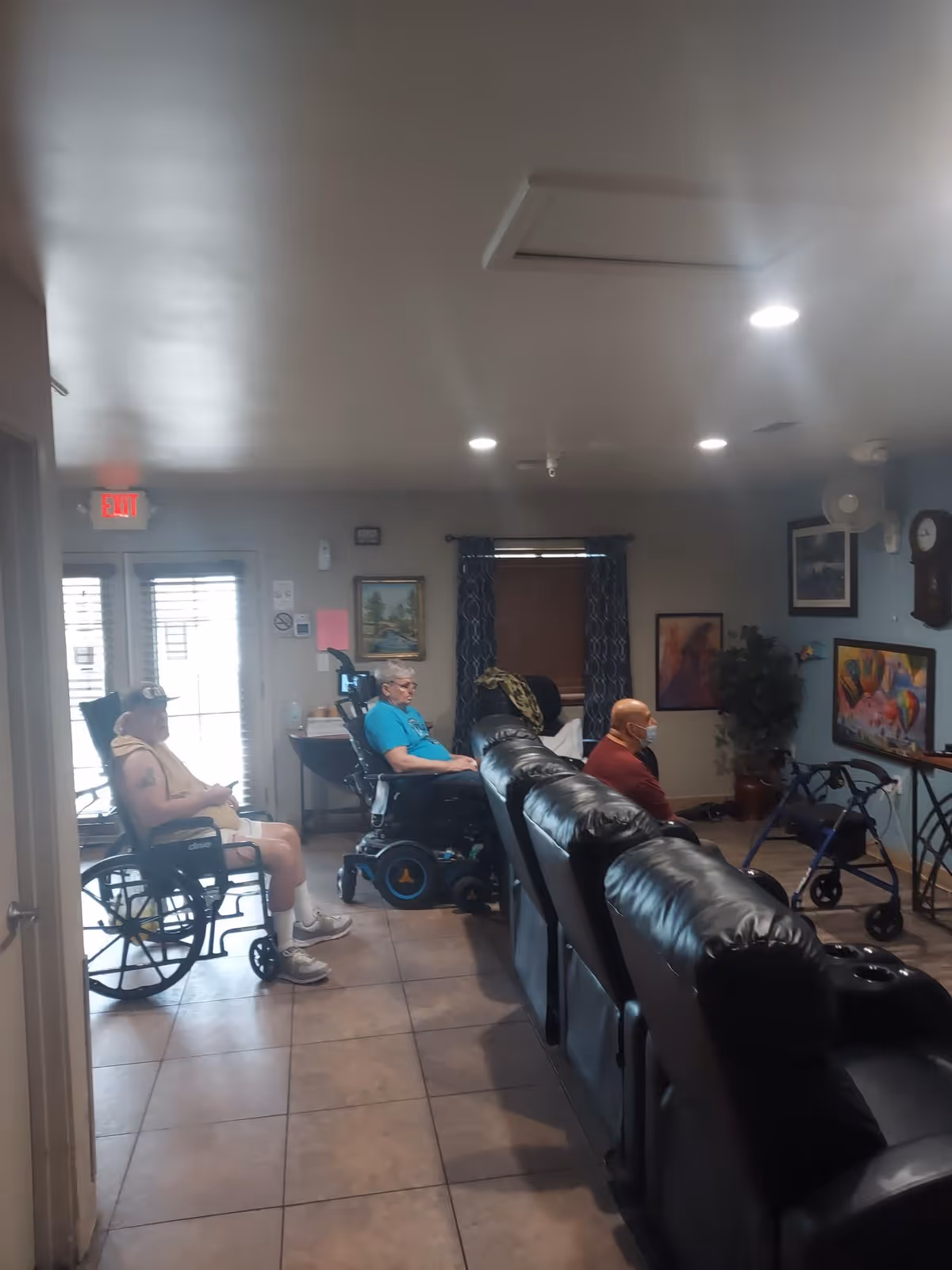 Three elderly individuals sitting in a living room area of a senior living facility. Two are in wheelchairs and one is seated on a black leather recliner. The room has tiled floors, a window with curtains, framed artwork on the walls, and a TV mounted on the wall. A walker is visible near the TV.