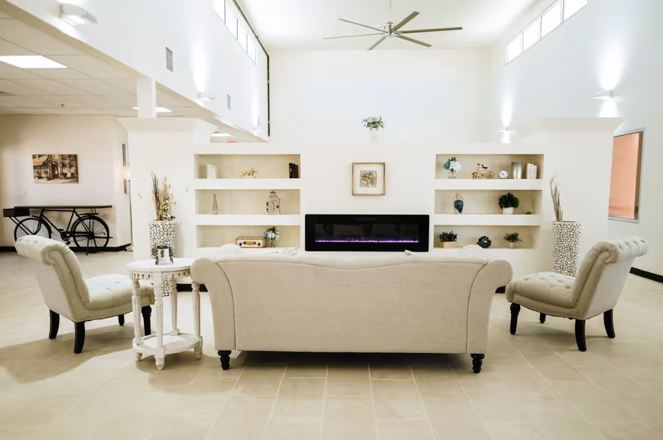 A bright and spacious living room with a beige sofa and two matching armchairs arranged around a modern electric fireplace set into a white built-in shelving unit. The shelves display decorative items including plants, vases, and small sculptures. The room features high ceilings with a large ceiling fan, tiled floors, and soft wall lighting. A small round side table is placed next to the sofa, and a bicycle is visible in the background near a wall with artwork.