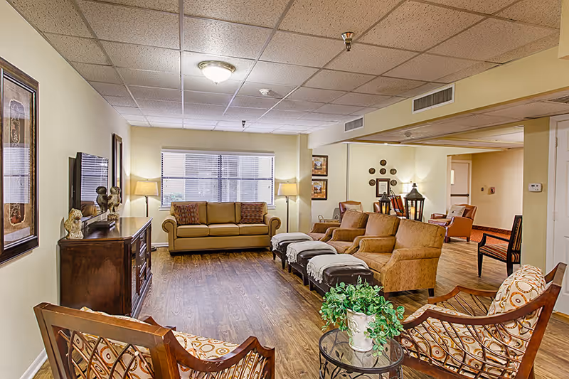 Bright, warmly furnished common living area with sofas, armchairs, ottomans, a TV cabinet and wood flooring.