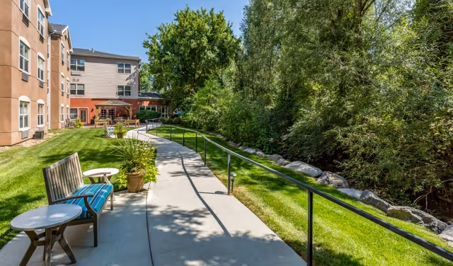 Outdoor walkway at Valencia at Cottonwood Heights with benches and small tables along a paved path. The path is bordered by a grassy area with plants and trees on one side and a building on the other. The scene is sunny with clear blue skies.