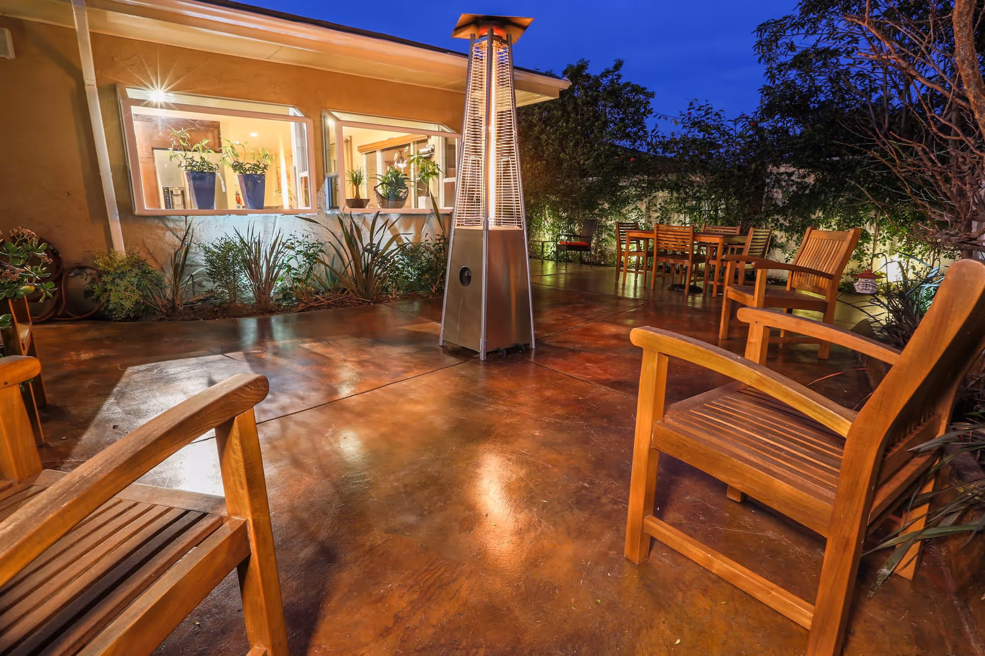 Outdoor patio area at dusk with wooden chairs and tables arranged around a tall outdoor heater. The patio has a polished concrete floor and is surrounded by plants and trees. The building wall has windows with potted plants visible inside.