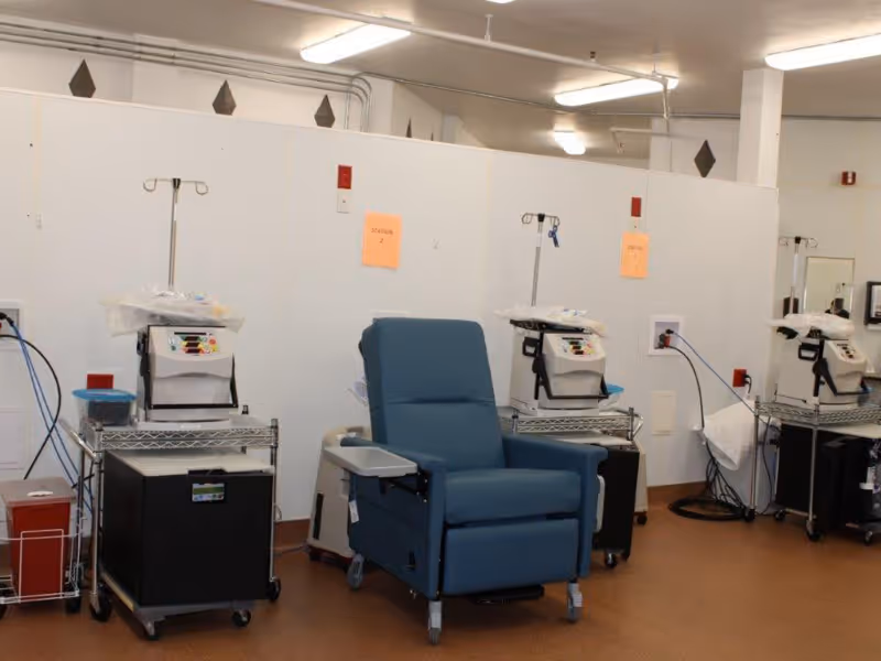 Medical treatment area with a blue reclining chair, IV poles, and medical machines on carts against a partitioned wall.