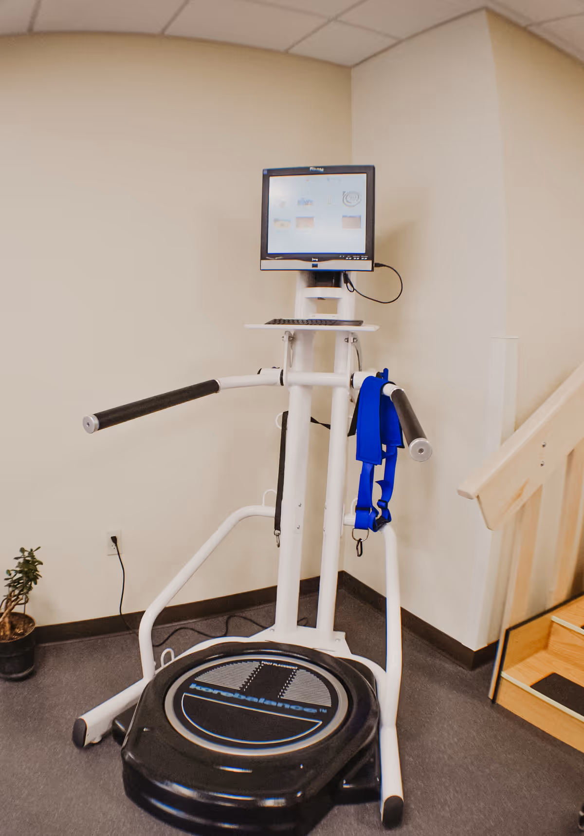 A vibration-platform exercise machine with handlebars, a mounted monitor, and a blue harness in a therapy room corner.
