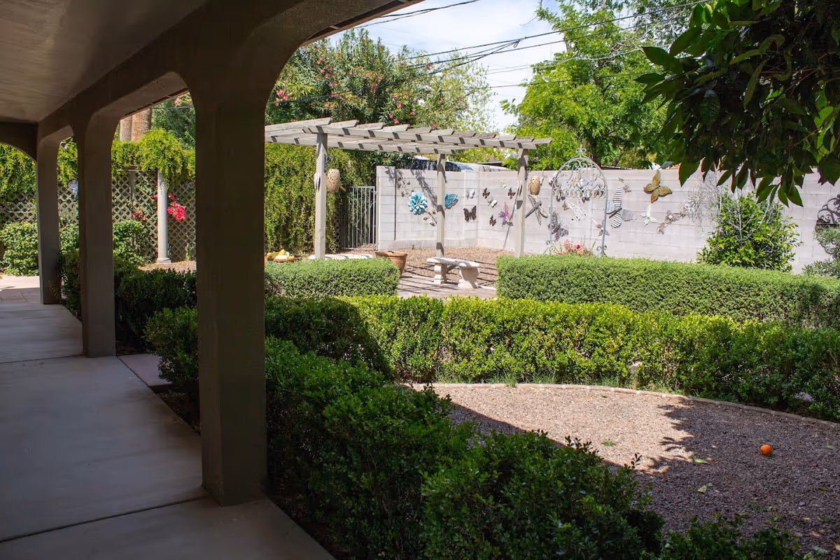 A shaded walkway with arches overlooks a garden area featuring trimmed hedges, a gravel path, a wooden pergola with a stone bench underneath, and a wall decorated with colorful butterfly ornaments. Trees and greenery surround the space under a clear sky.