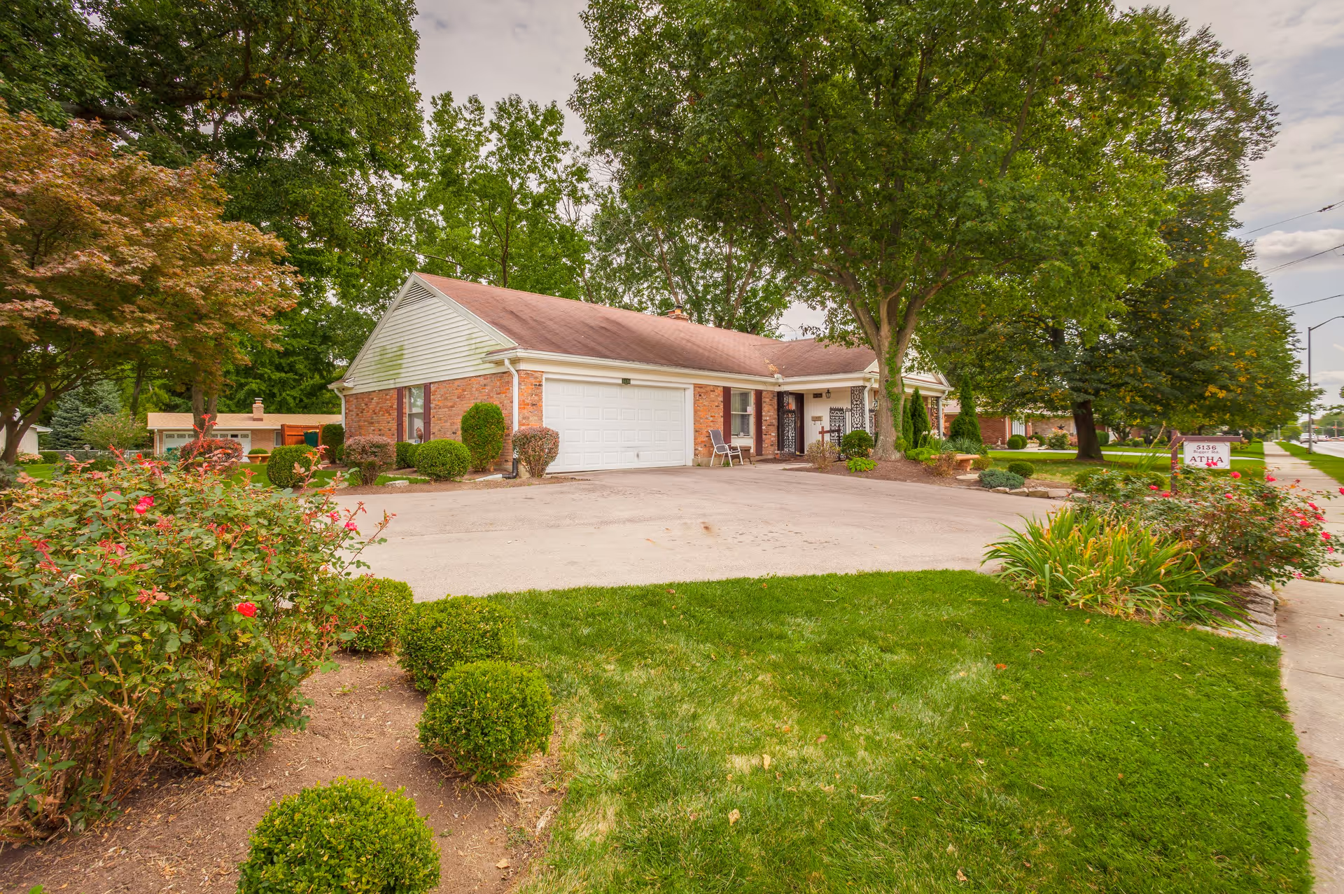 Exterior view of a single-story brick building with a white garage door, surrounded by green trees, bushes, and a well-maintained lawn. A paved driveway leads to the garage, and there is a sidewalk along the right side of the image. A sign near the sidewalk reads '5138 ATHA'.