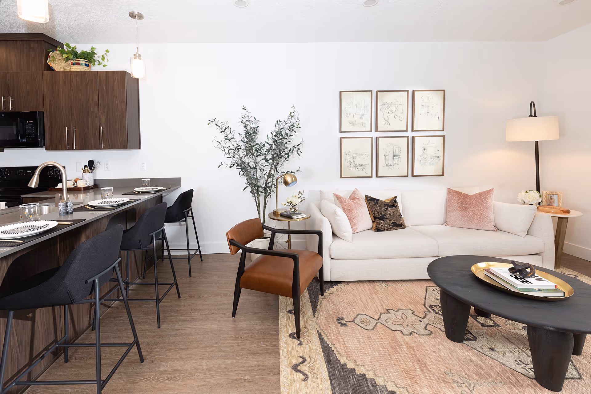 A modern open-concept living area featuring a kitchen with dark wood cabinets and a breakfast bar with four black bar stools. Adjacent to the kitchen is a cozy living room with a white sofa adorned with pink and black pillows, a brown leather armchair, a black oval coffee table with a gold tray and books, a floor lamp, and framed artwork on the white wall. The floor is wood with a patterned area rug.