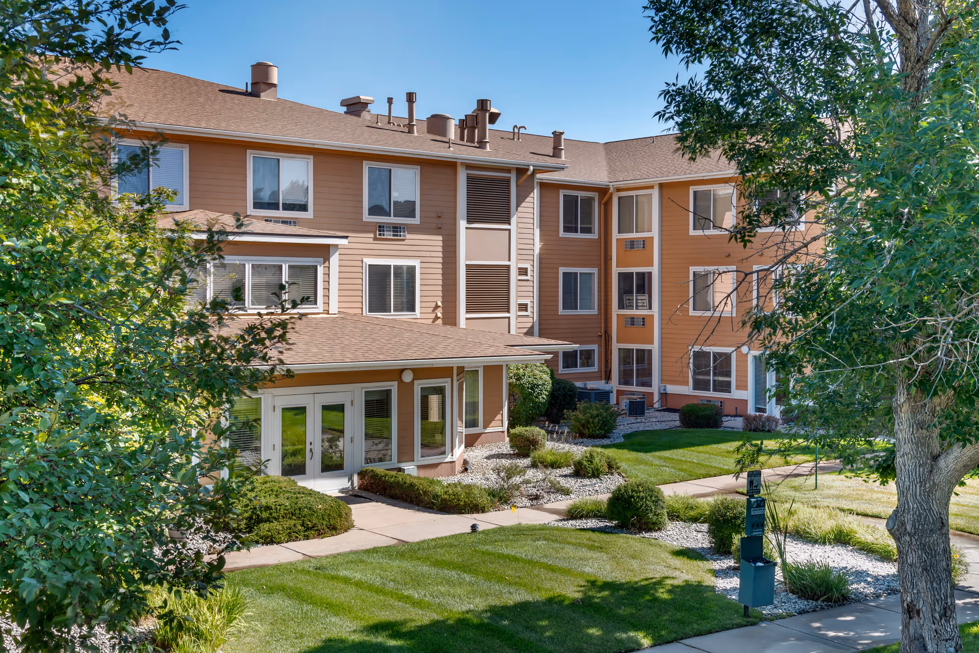Exterior view of a three-story senior living facility building with beige and brown siding, multiple windows, and a well-maintained green lawn with bushes and trees under a clear blue sky.