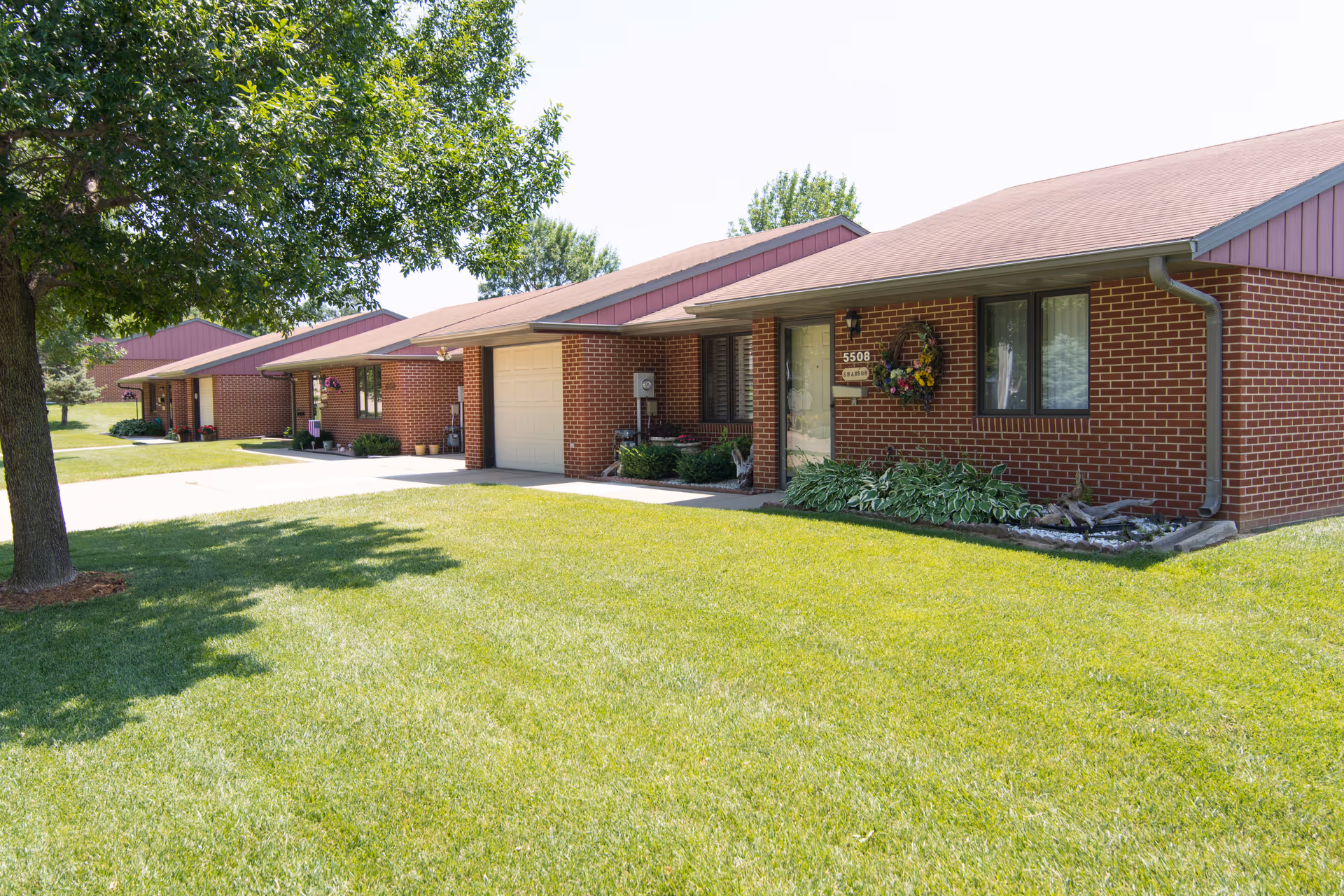 Exterior view of a single-story brick building with a sloped roof, a garage, and a front door decorated with a wreath. The building is part of a row of similar units with well-maintained green lawns and a large tree providing shade on the left side.