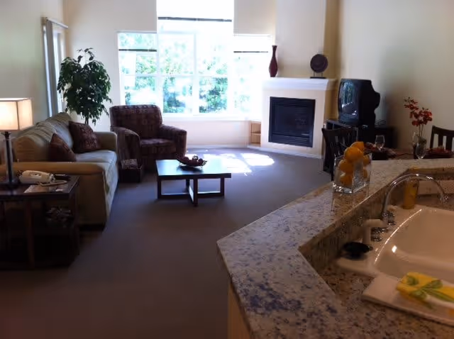 A cozy living room area with a beige sofa, a patterned armchair, a wooden coffee table, and a side table with a lamp. There is a large window letting in natural light, a fireplace with decorative items on the mantel, and a small TV on a stand. In the foreground, there is a kitchen counter with a sink, a glass container filled with oranges, and a folded towel with a lemon slice on it.