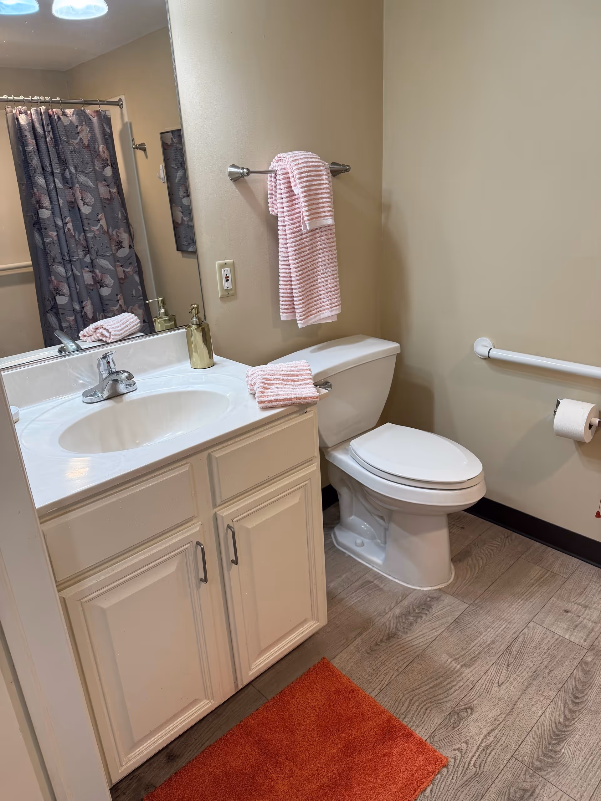 A bathroom with a white sink and cabinet, a toilet, a towel rack with a pink and white striped towel, a matching hand towel on the sink, a soap dispenser, a shower curtain with a floral pattern, and a red bath mat on the wood-patterned floor.