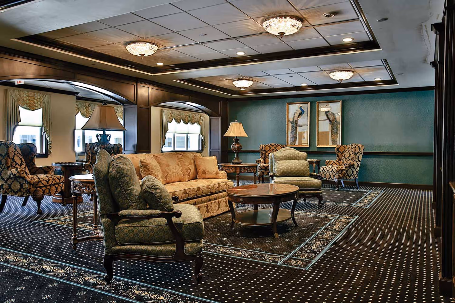 A spacious and elegantly decorated living room with patterned carpet, upholstered armchairs, a beige sofa, wooden coffee tables, table lamps, and framed artwork on the green walls. The ceiling features recessed lighting and chandeliers.