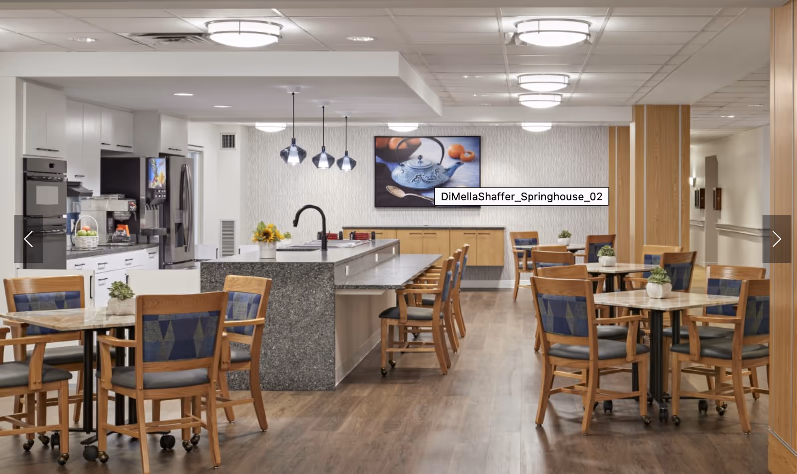 Bright communal dining area with multiple tables and chairs and a central kitchen island under ceiling lights.