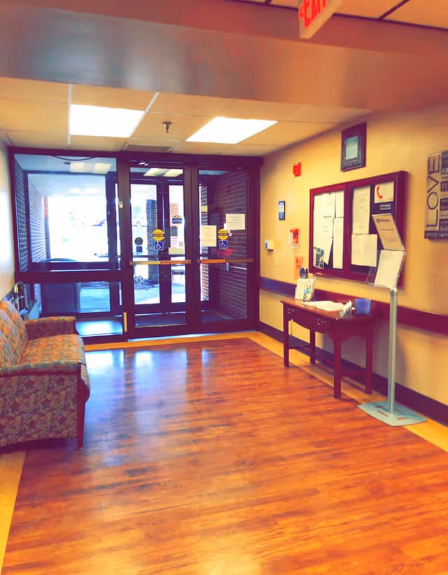 Interior view of a facility entrance area with wooden flooring, a floral patterned couch on the left, a small wooden table with hand sanitizer and papers on the right, bulletin boards on the wall, and glass double doors leading outside.