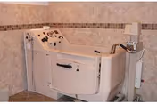 A specialized bathing tub designed for assisted bathing, featuring a door for easy access and control knobs, situated in a tiled room with beige walls and a small lamp on a stand nearby.