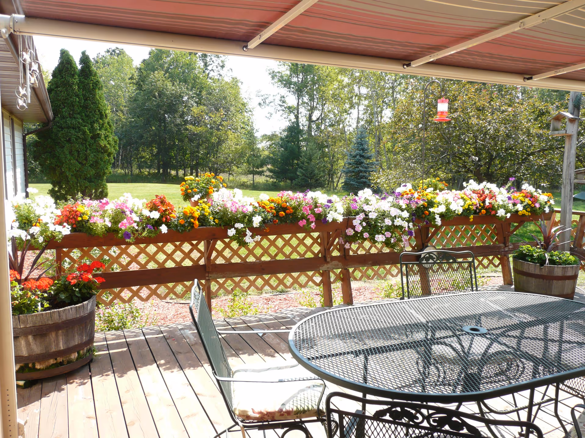 A wooden deck with a metal table and chairs under a retractable awning. The deck railing is decorated with colorful flower boxes filled with various blooming flowers. In the background, there is a green lawn and trees under a sunny sky.