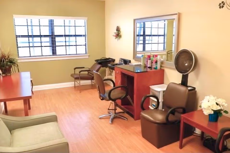Small salon-style interior with styling chairs, a mirror, a hooded hair dryer, and seating in a bright room.