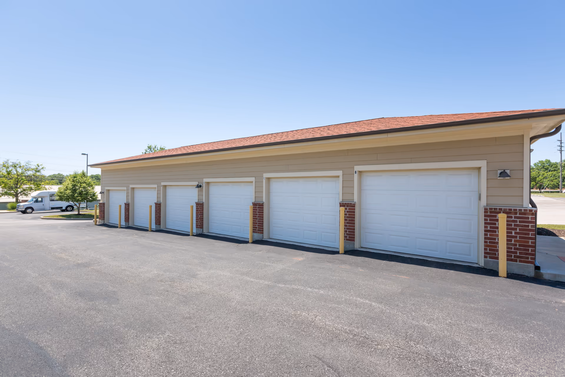 A row of six closed white garage doors on a beige building with a red roof, situated in a paved parking area under a clear blue sky. There are yellow bollards in front of some garage doors and a white truck is parked in the background near some trees.