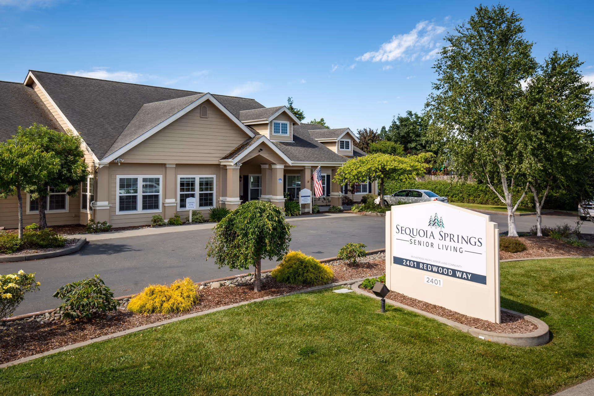 Exterior view of Sequoia Springs Senior Living Community building with beige siding, multiple windows, and a peaked roof. The entrance has a small covered porch with columns and an American flag. The foreground features a landscaped area with green grass, small trees, and shrubs. A large sign in front displays the facility name and address: 2401 Redwood Way.