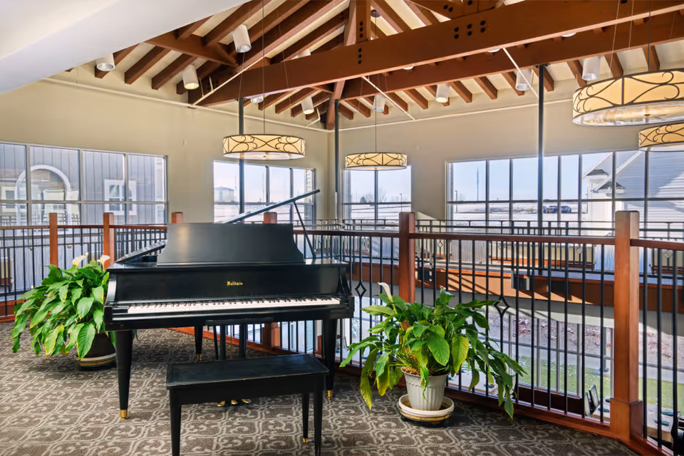 Interior view of a senior living facility area with a black grand piano and matching bench placed on a patterned carpet. The space features large windows allowing natural light to fill the room, wooden beams on the ceiling, decorative hanging light fixtures, and two green potted plants on either side of the piano. A railing surrounds the area, overlooking a lower level.