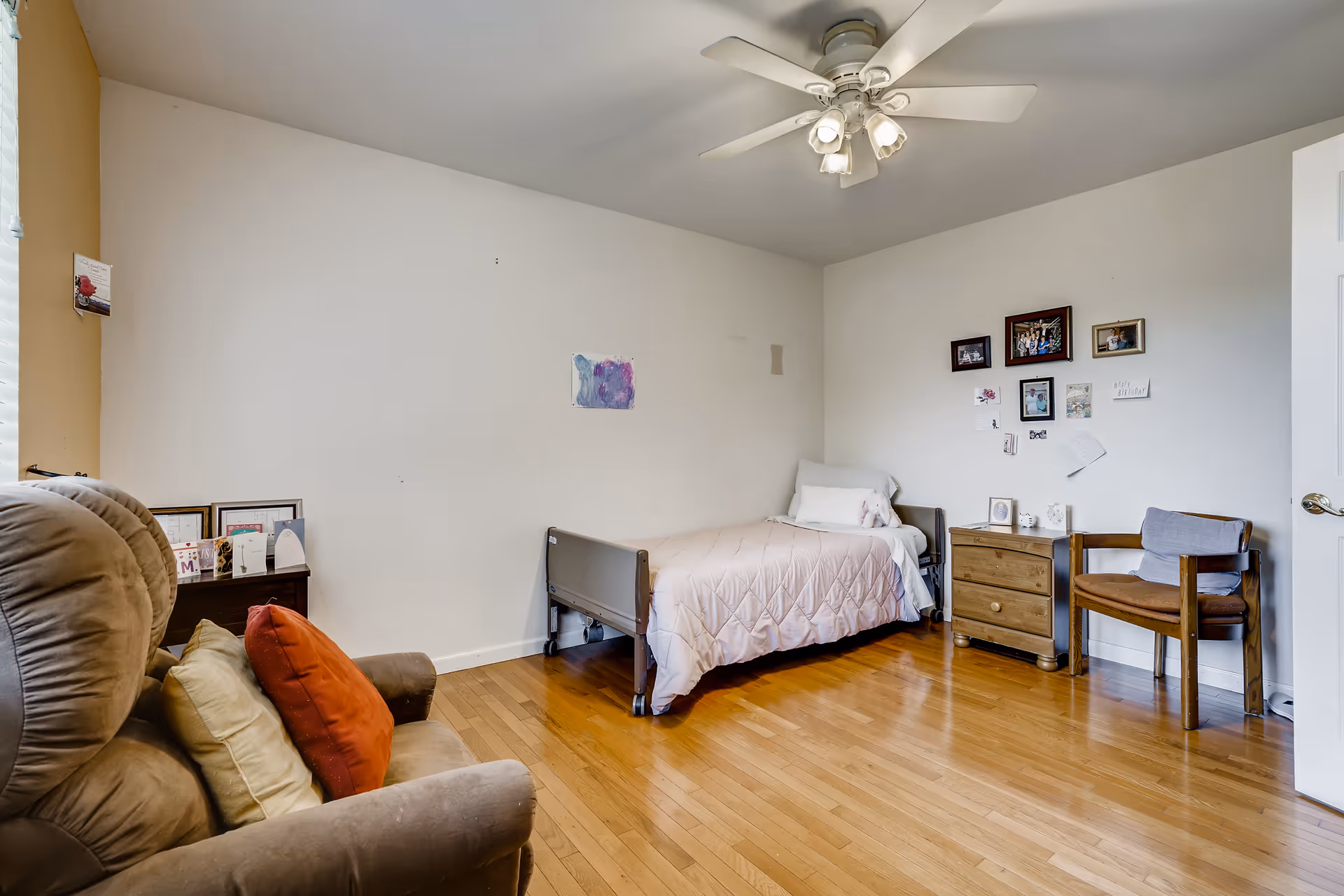 Simple bedroom with a twin bed, nightstand, armchair and wooden floor under a ceiling fan.