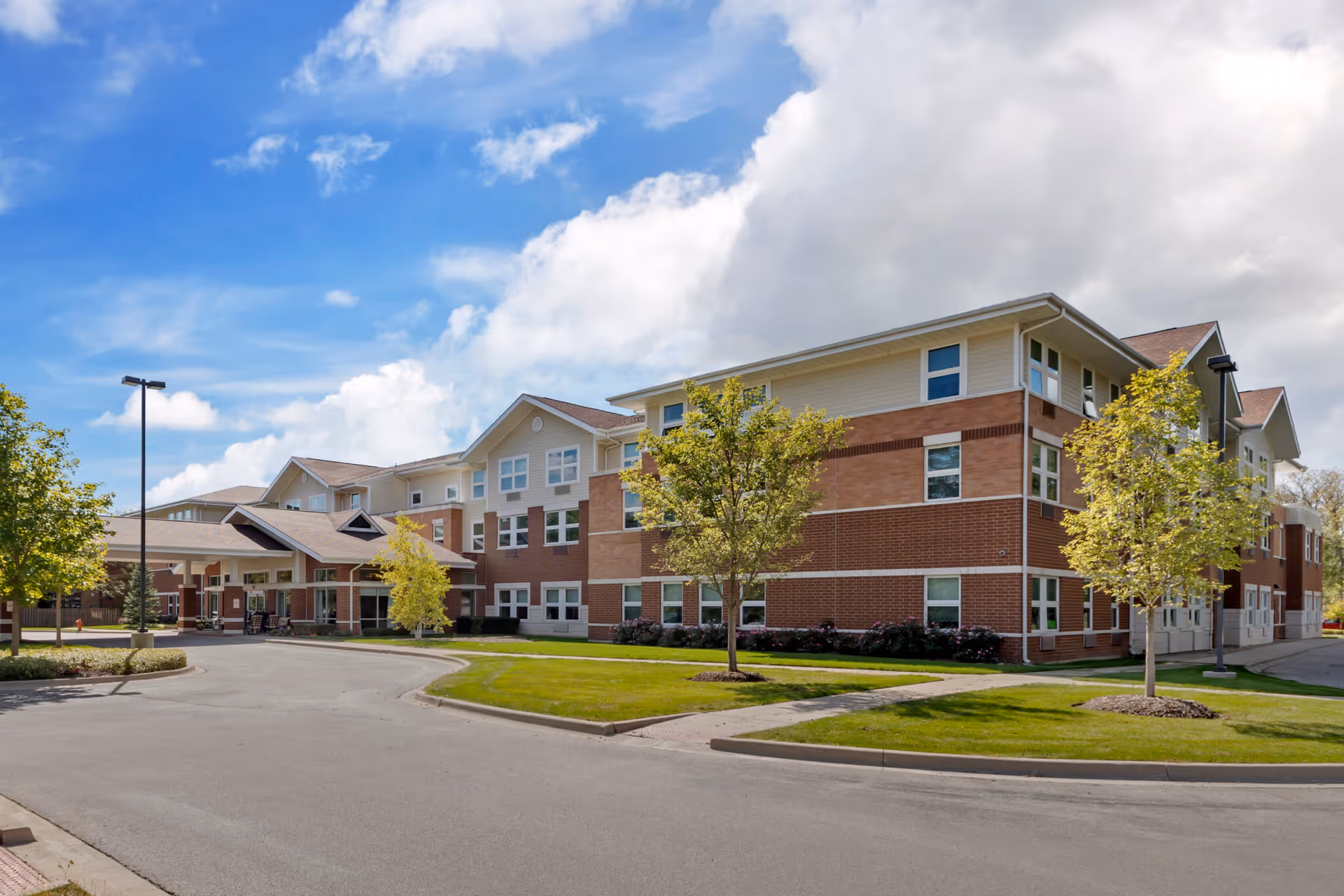 Exterior view of a three-story senior living facility building with a covered entrance, surrounded by green lawns and trees under a partly cloudy blue sky.