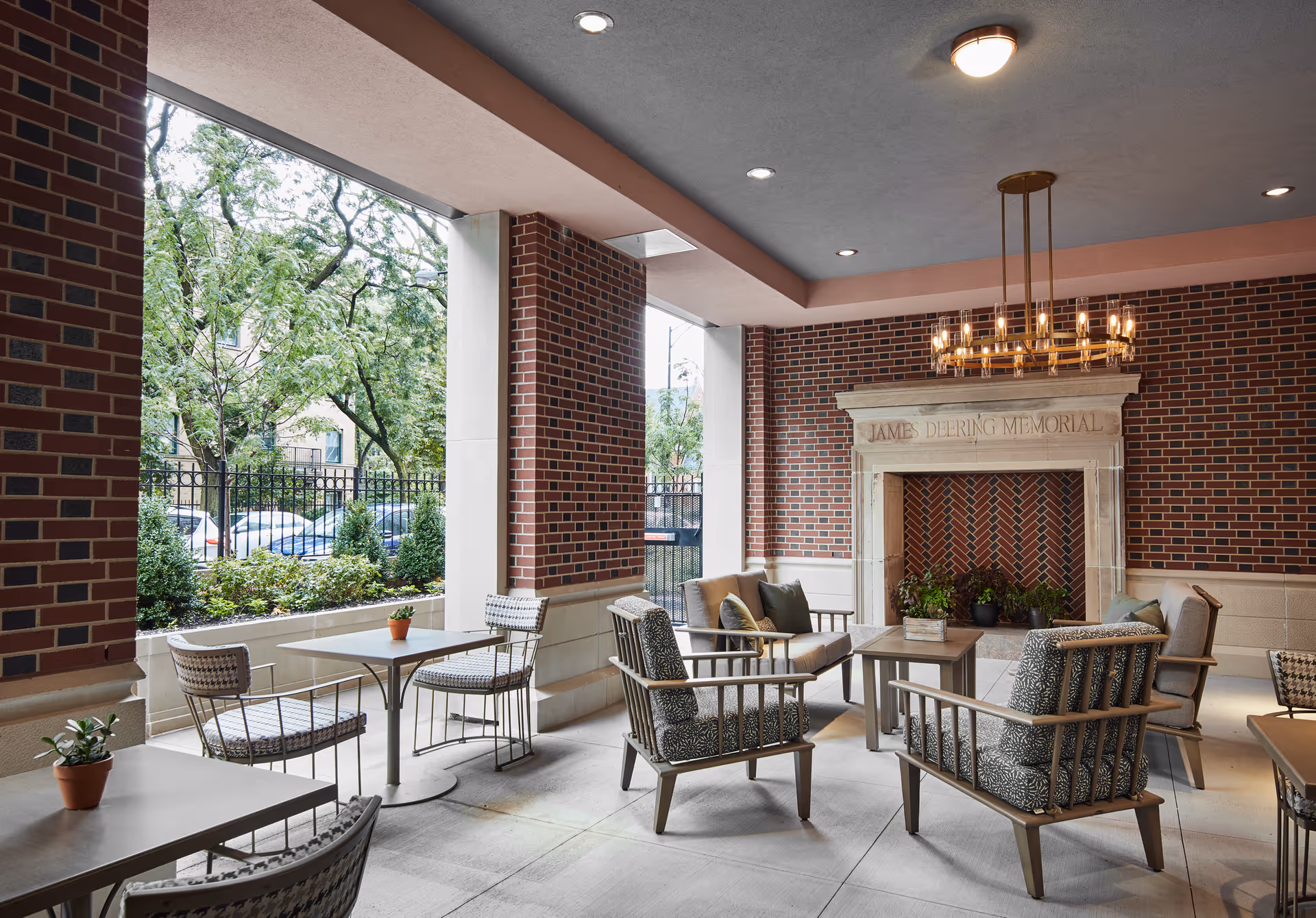 Covered outdoor seating patio with tables, chairs, potted plants, and a decorative fireplace set against brick walls.