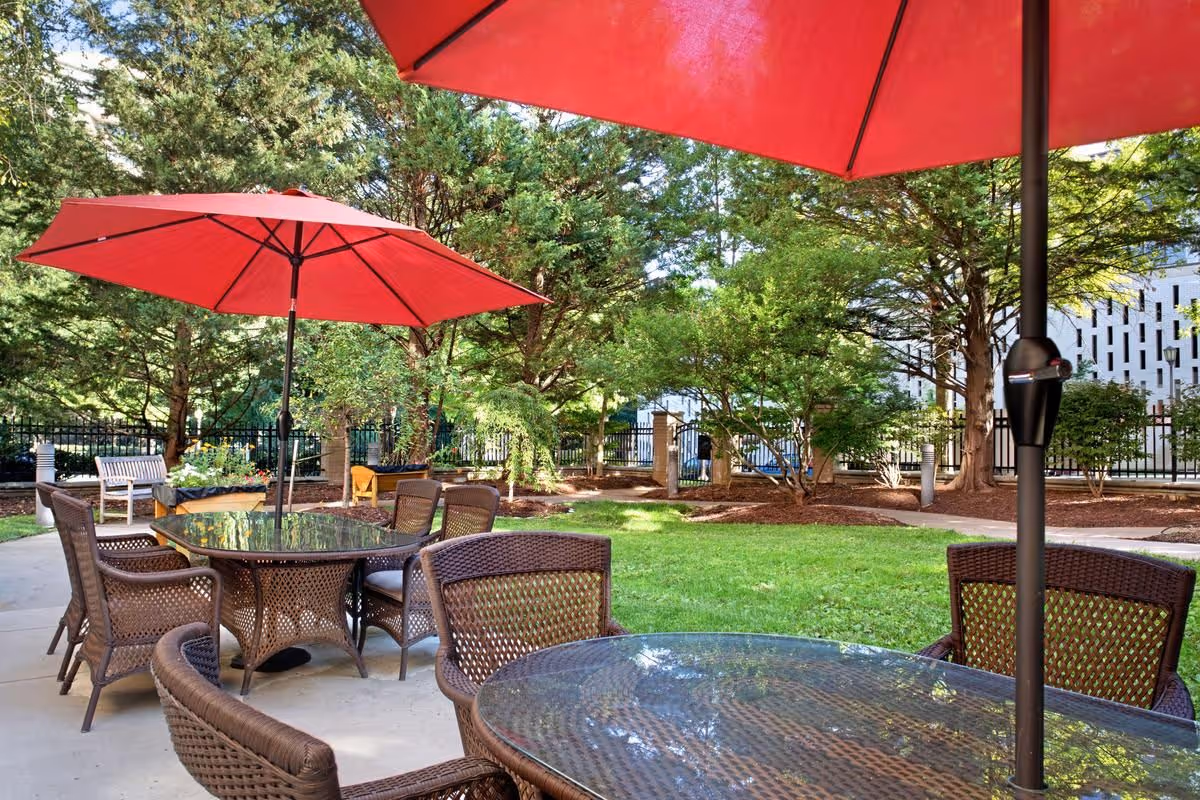 Outdoor patio area with glass-top tables and brown wicker chairs under large red umbrellas, surrounded by green grass, trees, and a black metal fence in the background.
