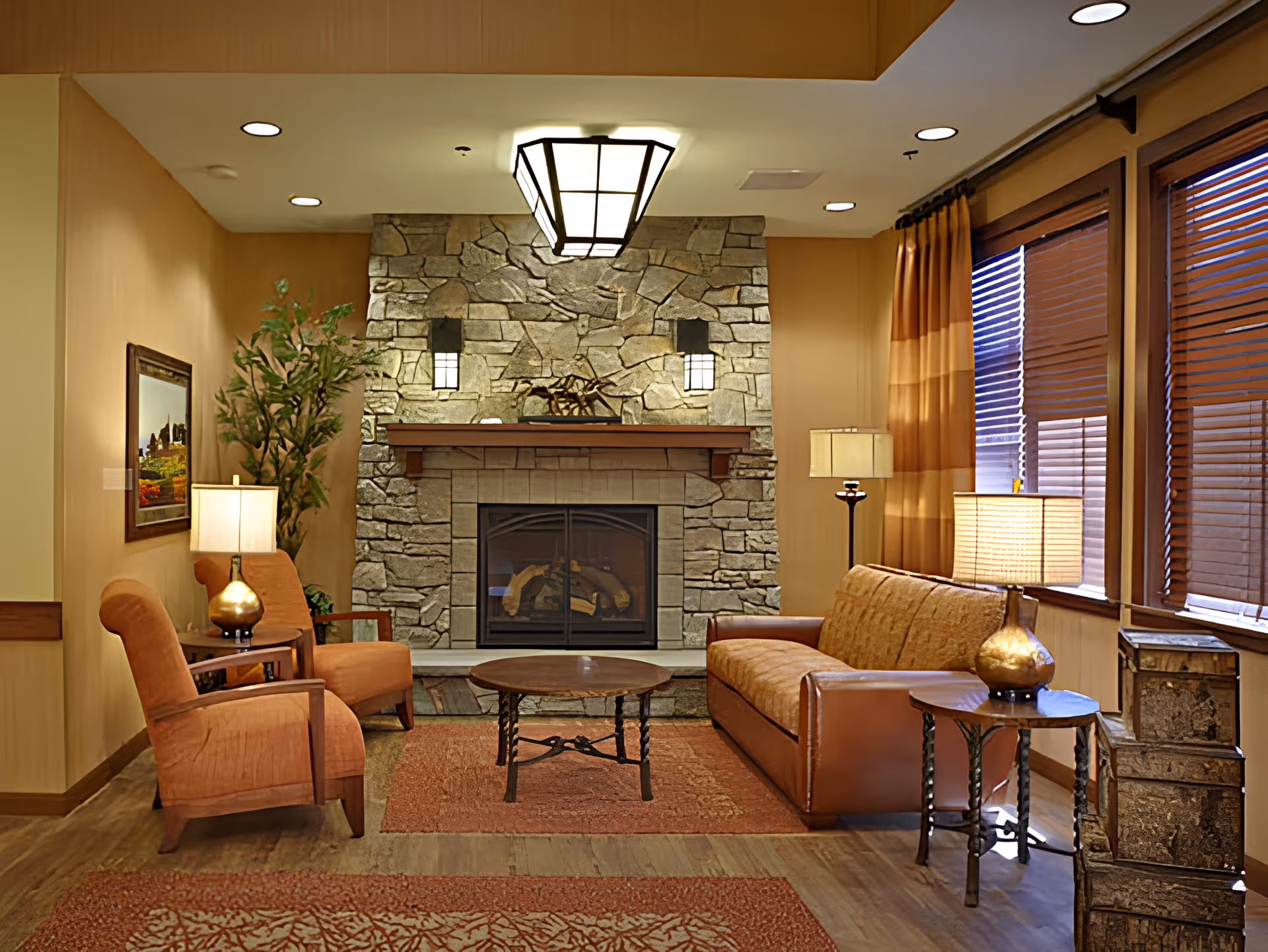 Cozy living room with a stone fireplace, sofa and armchairs, coffee table, lamps, and large windows with blinds.
