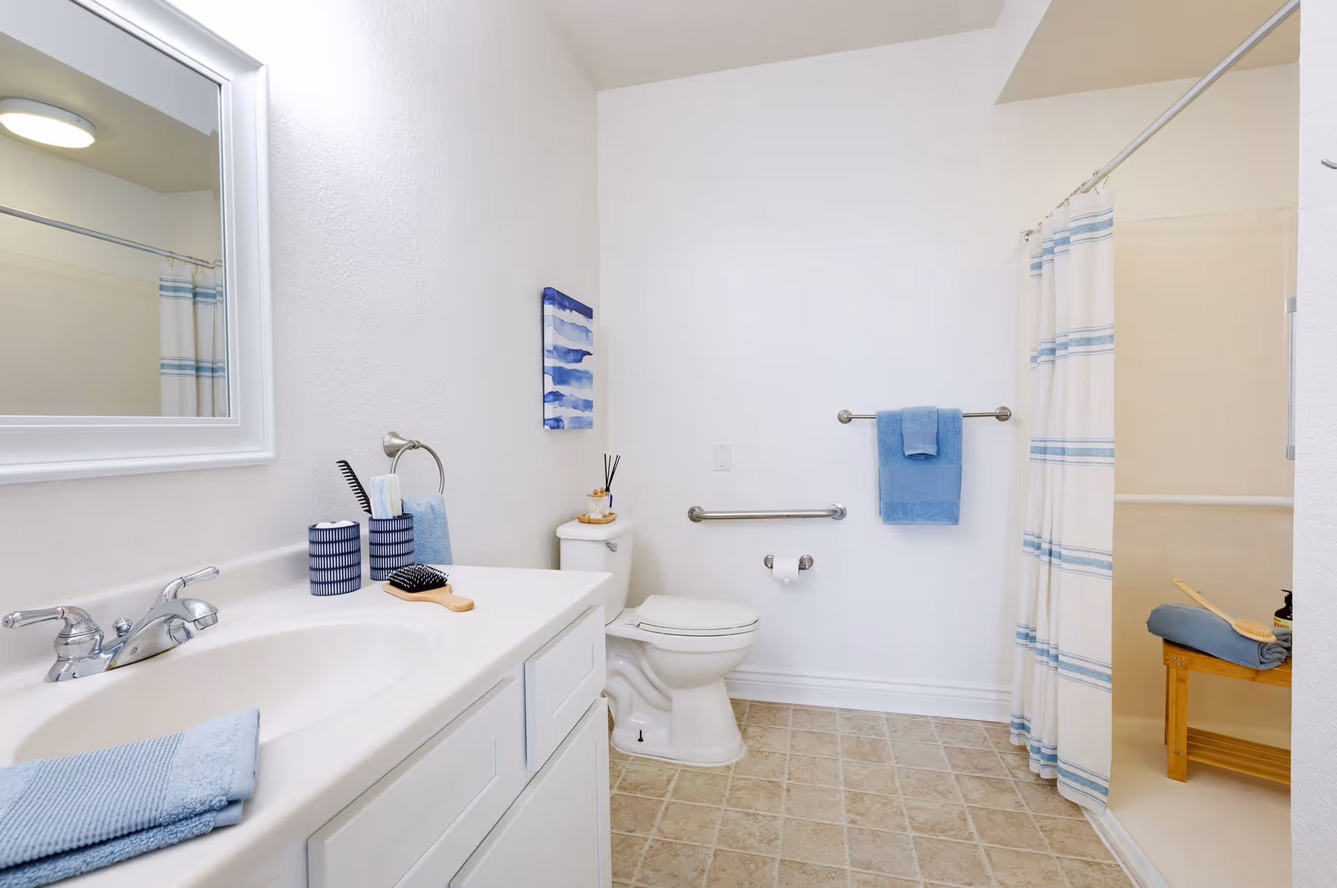 A clean and bright bathroom with a white sink and countertop on the left, a large mirror above the sink, a toilet in the center, and a shower with a curtain on the right. The bathroom features blue towels, a small wooden brush, and decorative items on the countertop and toilet tank. The floor is tiled, and there are grab bars near the toilet for accessibility.