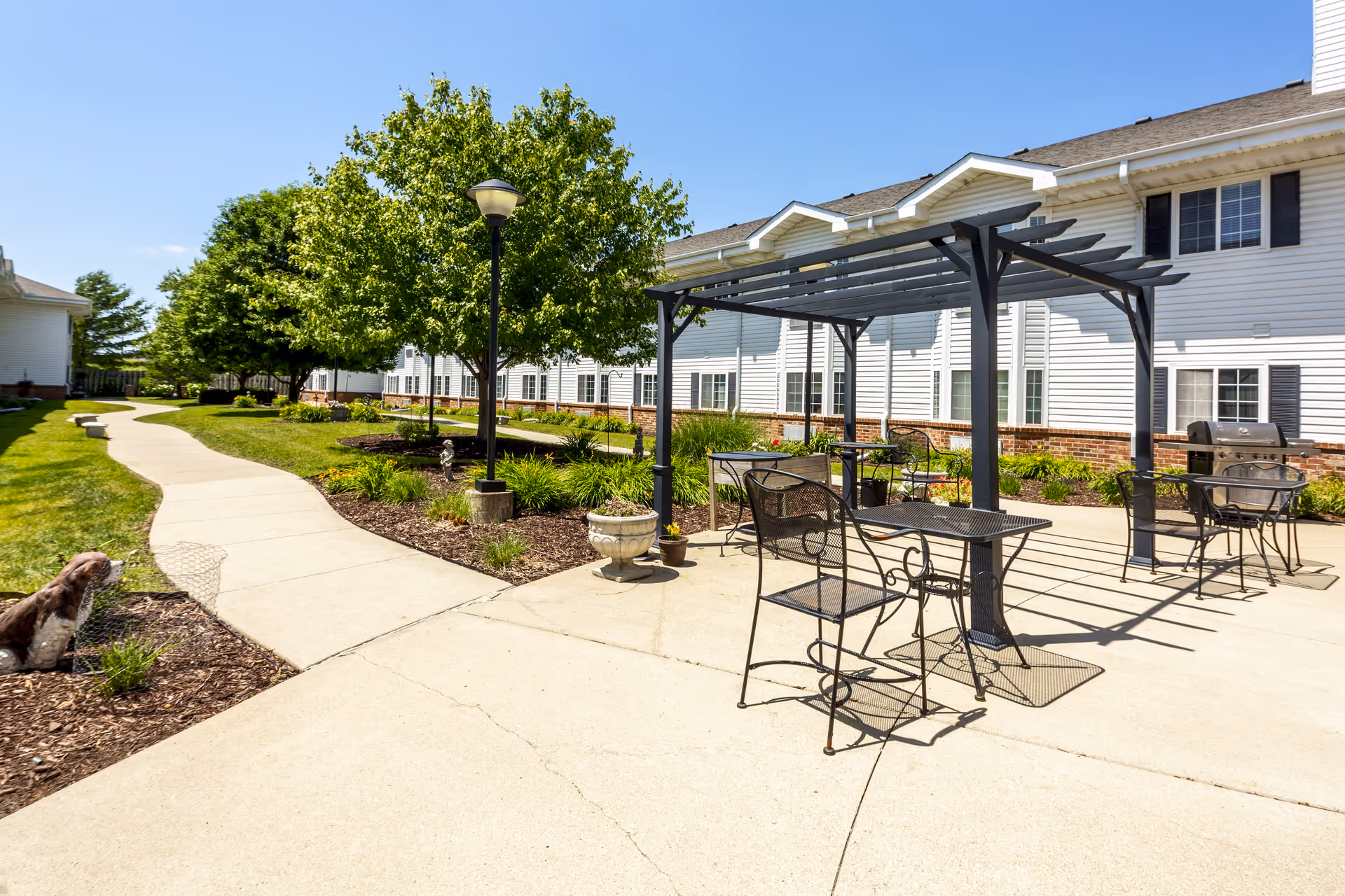 Outdoor patio area at Independence Village Ames featuring a paved walkway, metal tables and chairs under a pergola, a barbecue grill, green trees, and a two-story white building with black shutters in the background under a clear blue sky.