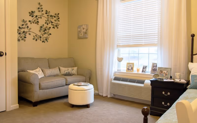A cozy living area with a gray loveseat adorned with decorative pillows, a small round white ottoman in front, and a window with white blinds and sheer curtains. There is a black nightstand with framed photos and a cup next to a bed with a blue pillow. A decorative metal wall art piece and a small floral painting hang on the beige walls.