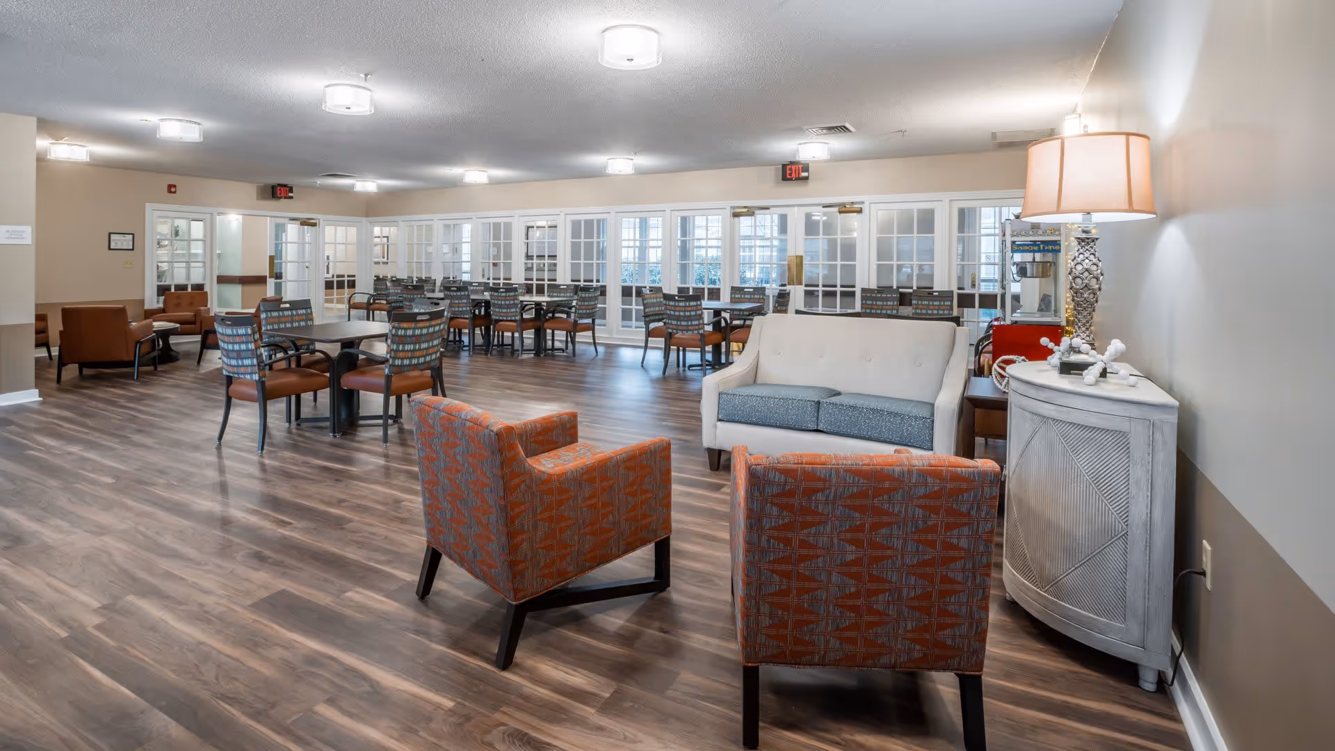 A spacious common area with wood flooring featuring multiple seating arrangements including patterned armchairs, a white loveseat, and several tables with chairs. The room is well-lit with ceiling lights and has large windows and glass doors along one wall. A small side table with a lamp and decorative items is positioned against the right wall.