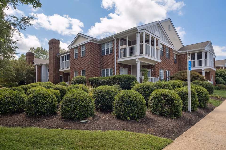 Exterior view of a two-story brick building with white balconies and multiple windows, surrounded by neatly trimmed bushes and a sidewalk. The sky is partly cloudy.