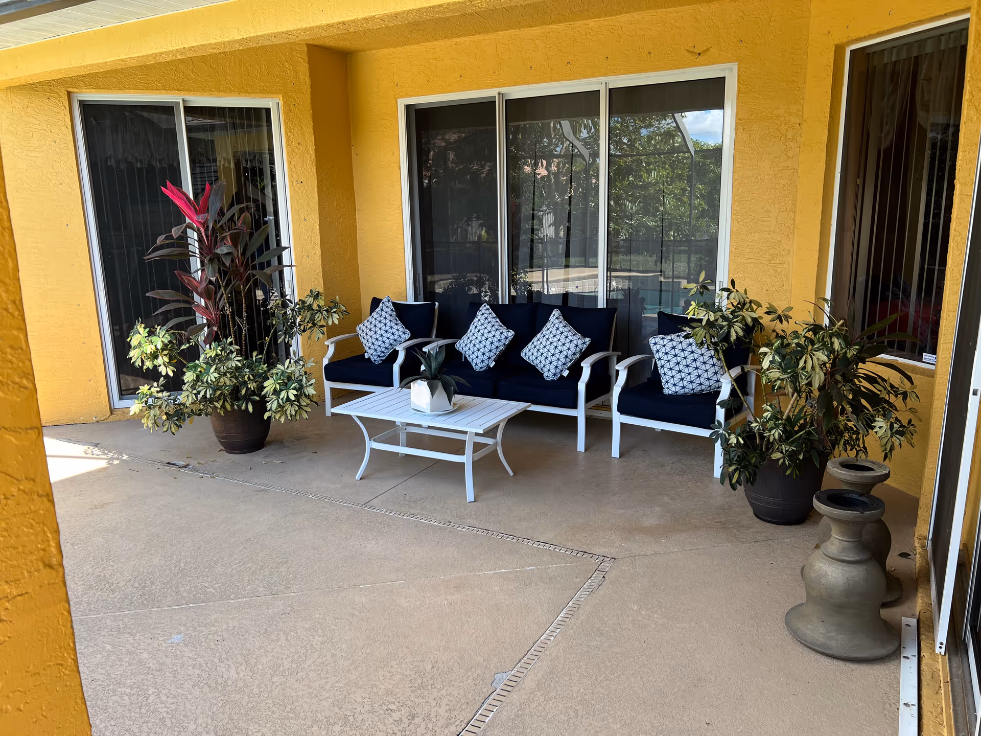 Outdoor patio area with yellow walls, featuring a white metal-framed sofa and two chairs with dark blue cushions and patterned throw pillows. A white coffee table with a small potted plant is in front of the seating. There are two large potted plants on either side of the seating area, and sliding glass doors behind the furniture.