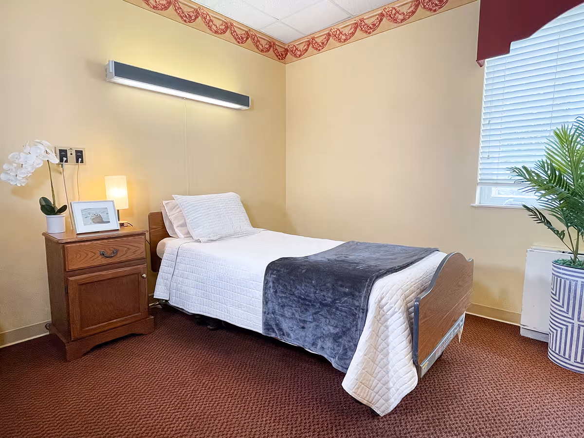 A small, neatly made single bed with white bedding and a dark gray blanket folded at the foot. Next to the bed is a wooden nightstand with a white orchid plant, a framed photo, and a small lamp. The room has beige walls with a decorative border near the ceiling, a window with white blinds and a red valance, and a potted plant in a blue and white striped pot on the floor.