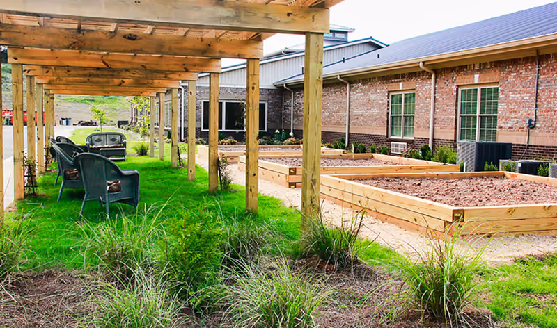 Outdoor garden area at Thomas Kelly Senior Living Community featuring raised garden beds with soil, a wooden pergola with seating including chairs and a bench, green grass, and plants. The background shows a brick building with windows.
