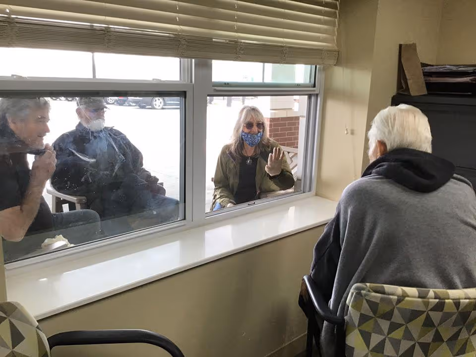 An elderly man sits inside a room facing a window, while three people sit outside on the other side of the window. One of the people outside, a woman wearing a face mask and sunglasses, is waving through the glass. The scene suggests a visit with physical separation for safety.