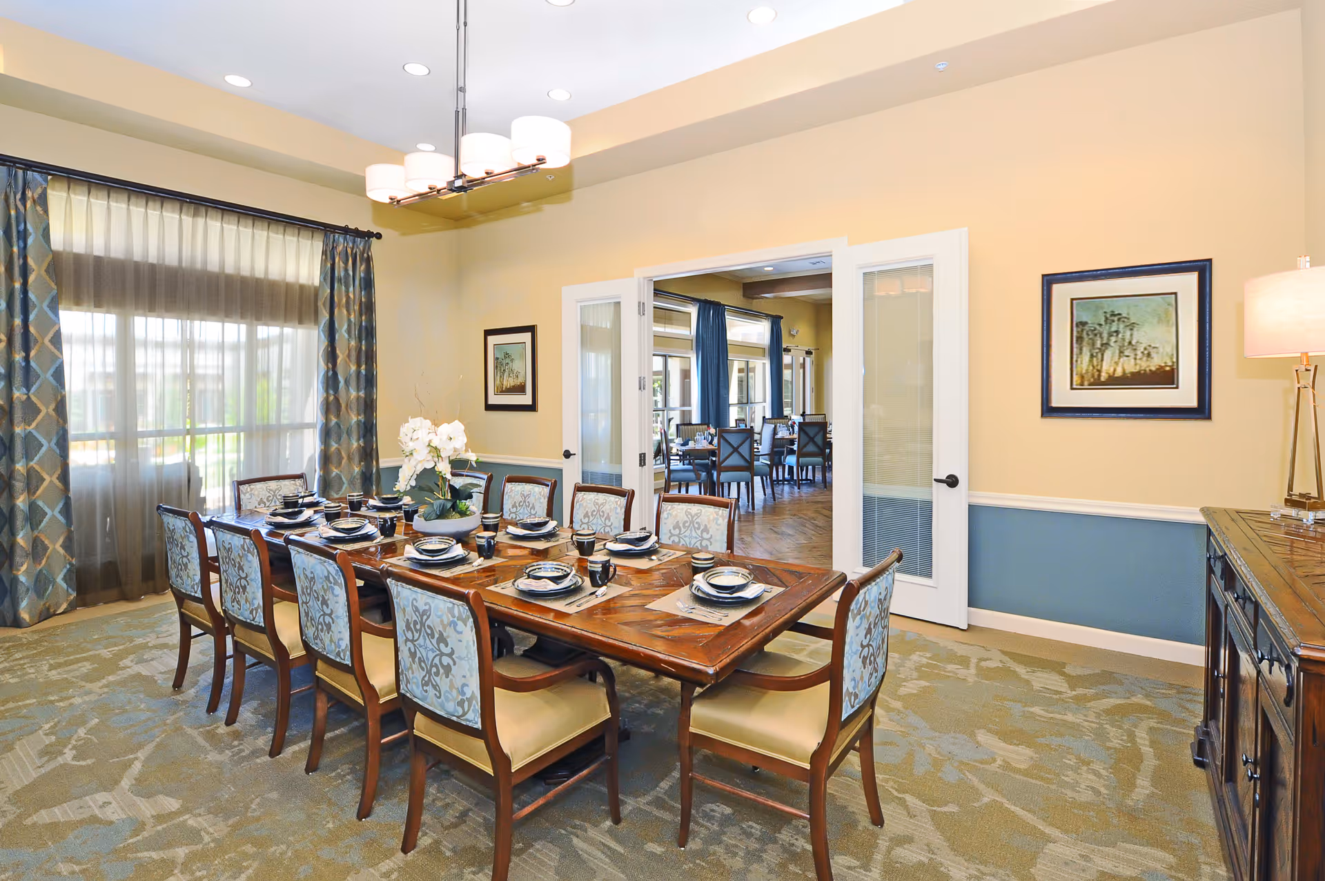 A well-lit dining room with a long wooden table set for eight people with plates, bowls, and cups. The chairs have blue patterned upholstery and cream-colored cushions. There are large windows with blue and gold patterned curtains allowing natural light to enter. A modern chandelier hangs above the table. The room has light yellow walls with a blue wainscot and framed artwork. Double glass doors open to another dining area with more tables and chairs.