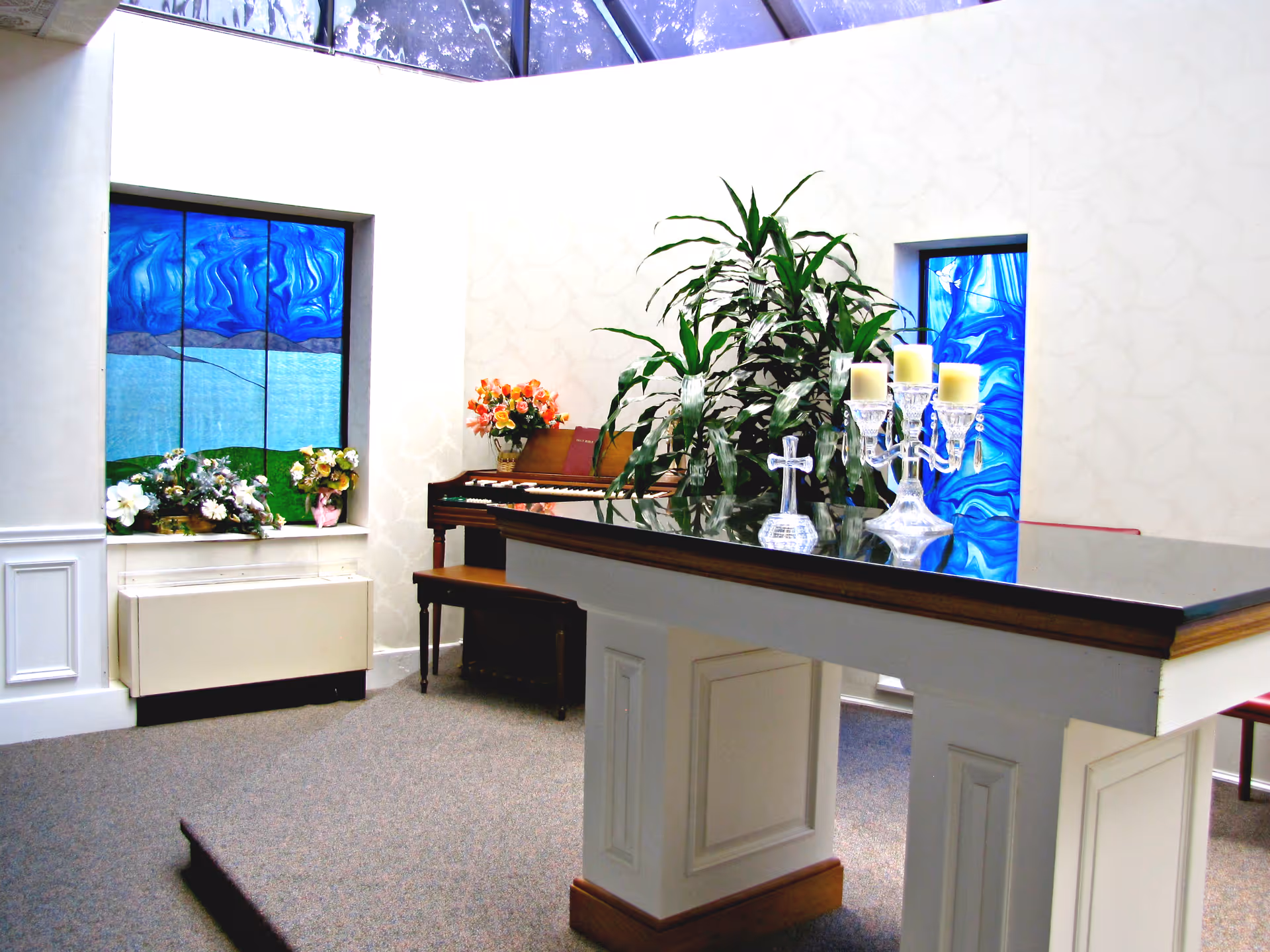 A bright room with stained glass windows featuring blue and green designs. There is a piano with a bench, a large green plant, and a white altar-like table with a glass cross and a crystal candle holder with three candles. Floral arrangements are placed near the windows.