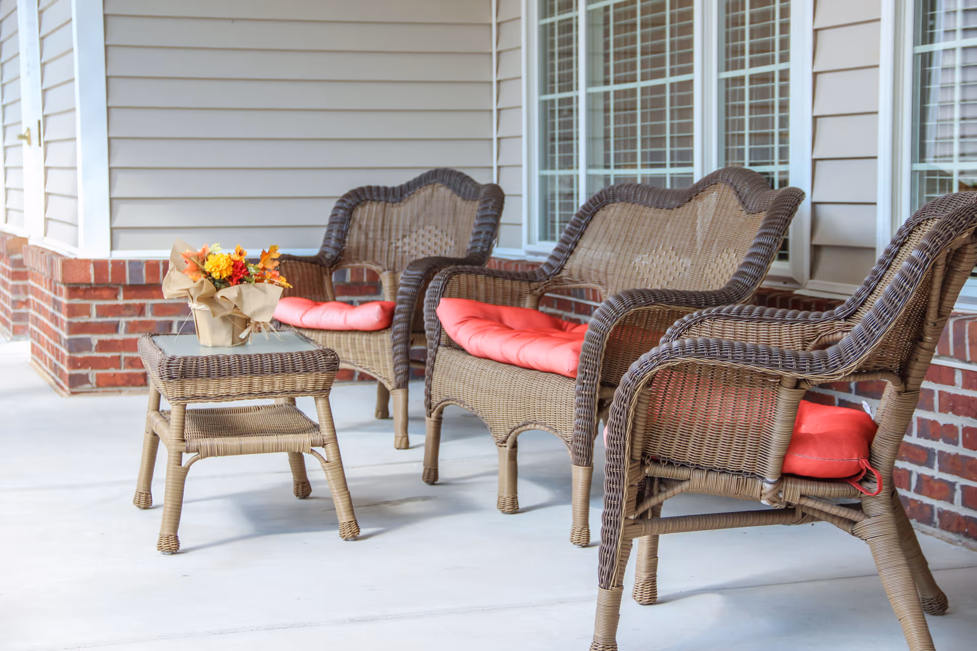 A porch area with three wicker chairs and a wicker table. The chairs have red cushions, and the table has a small flower arrangement with autumn-colored flowers. The porch has a brick base and beige siding with windows in the background.