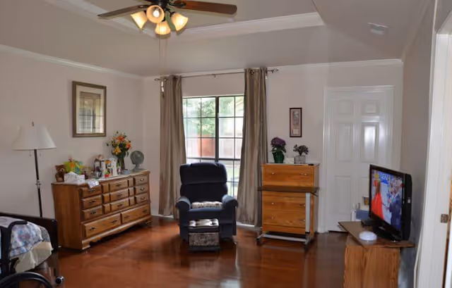 A cozy living room with wooden flooring, a ceiling fan with lights, a large window with curtains, a dark blue recliner chair with a footrest, a wooden dresser with flowers and decorative items, a wooden cabinet with plants on top, and a TV on a wooden stand displaying a program.
