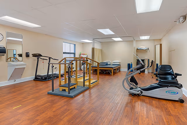 Physical therapy and exercise room with treadmills, a recumbent bike, parallel bars, and treatment tables in a senior living facility.