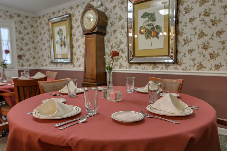 A dining room table set for four with a red tablecloth, white napkins folded on plates, drinking glasses, and silverware. The background features floral wallpaper, framed botanical prints, a tall wooden grandfather clock, and a vase with a single red rose on the table.