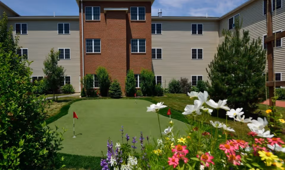 Outdoor putting green with small red flags surrounded by green grass, colorful flowers, and shrubs in front of a multi-story residential building with beige siding and red brick accents under a partly cloudy sky.