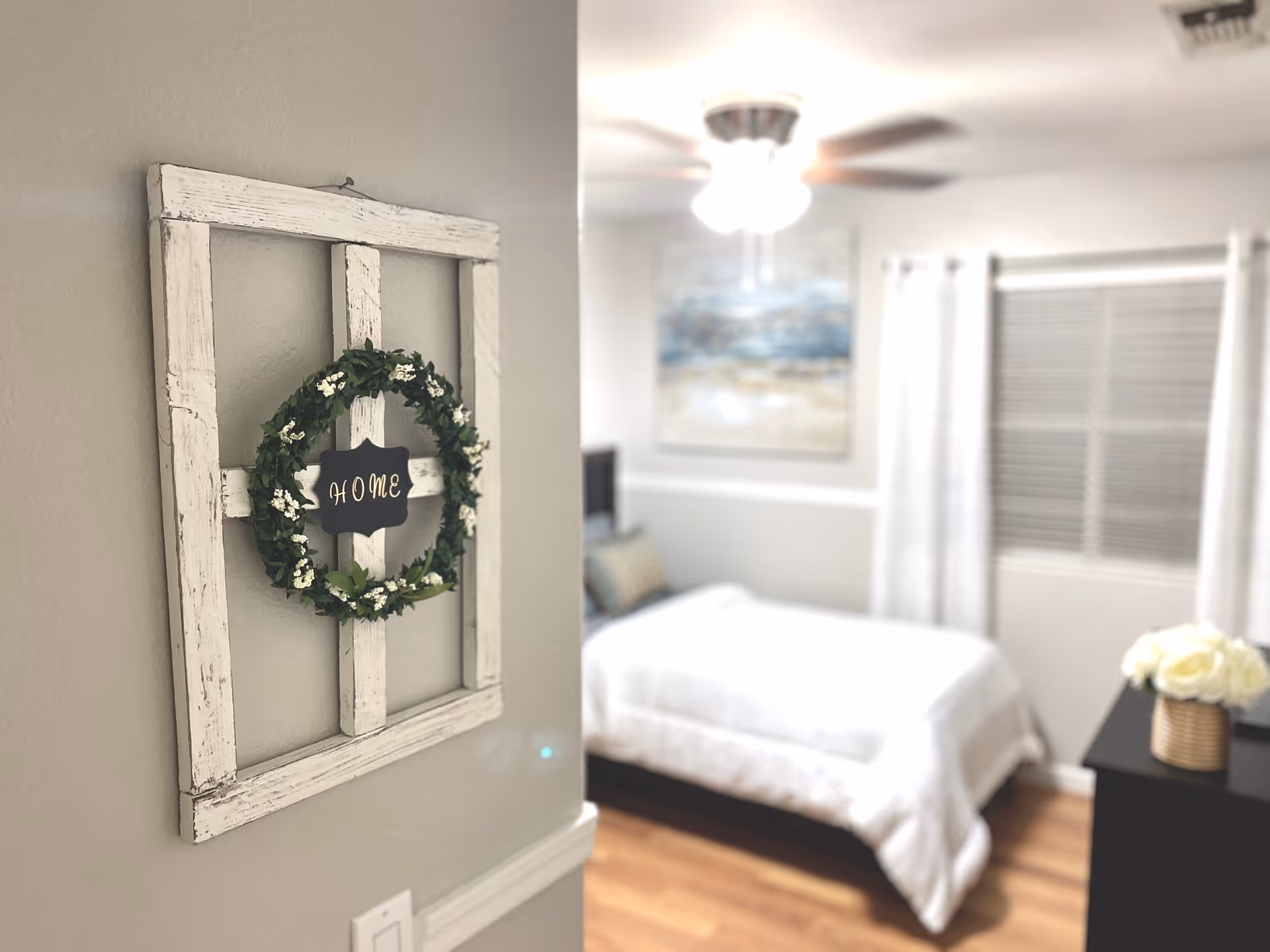 A cozy bedroom with a white bedspread, a ceiling fan with lights, a window with white curtains, and a black dresser with a vase of white flowers. On the wall near the entrance, there is a rustic white wooden frame decorated with a green wreath and a small sign that says 'HOME'.