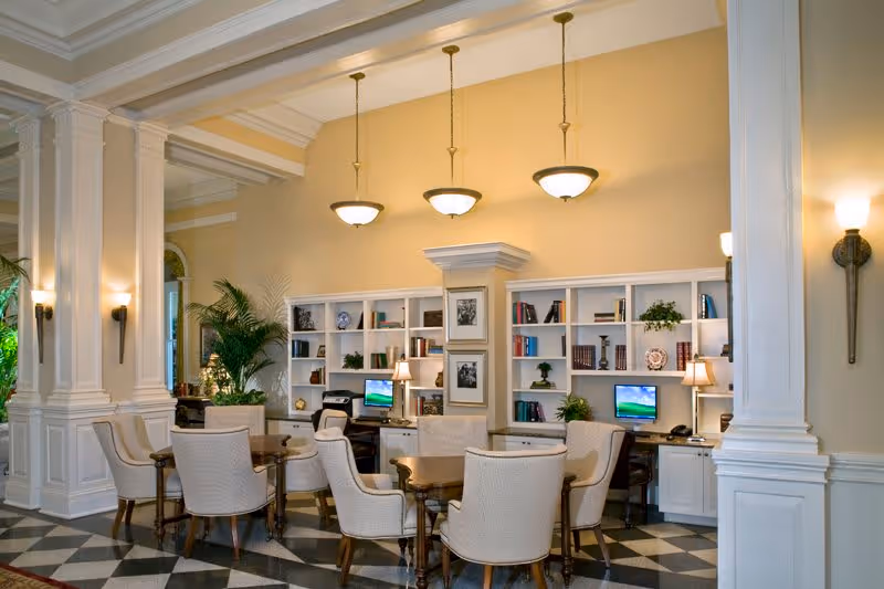 A spacious, elegant common area with cream-colored walls and high ceilings featuring three hanging pendant lights. The room has white built-in bookshelves filled with books, plants, and decorative items. There are two computer desks with desktop computers and table lamps. Several upholstered armchairs surround wooden tables, and large white columns frame the space. The floor has a black and white checkered pattern.